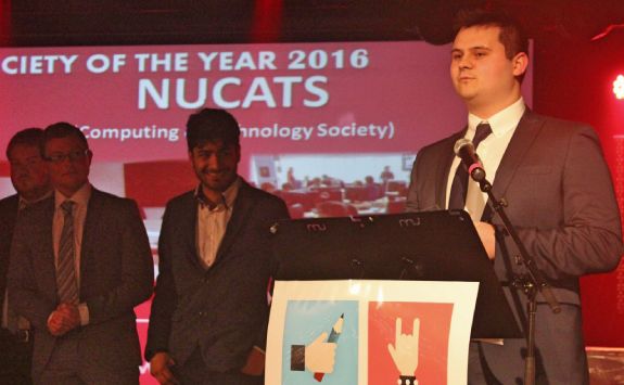A photo of the 2016 NUCATS president giving a speech, the background has a photo saying Society of the Year 2016, NUCATS.